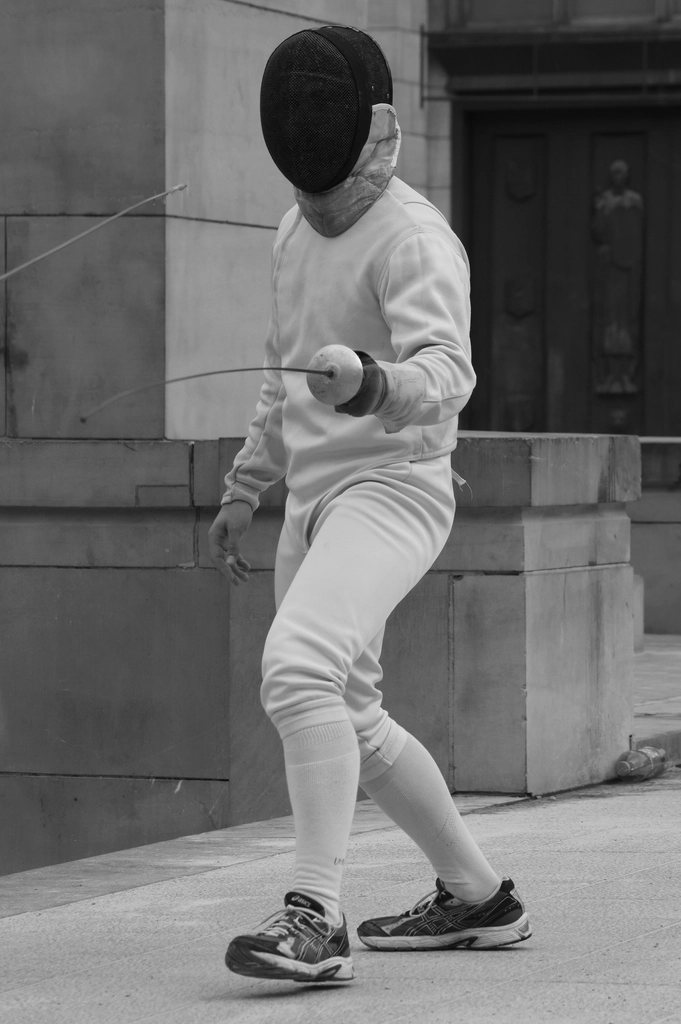 A fencer in a black mask and white jacket stands guard on a stone pavement, photographed in black and white.