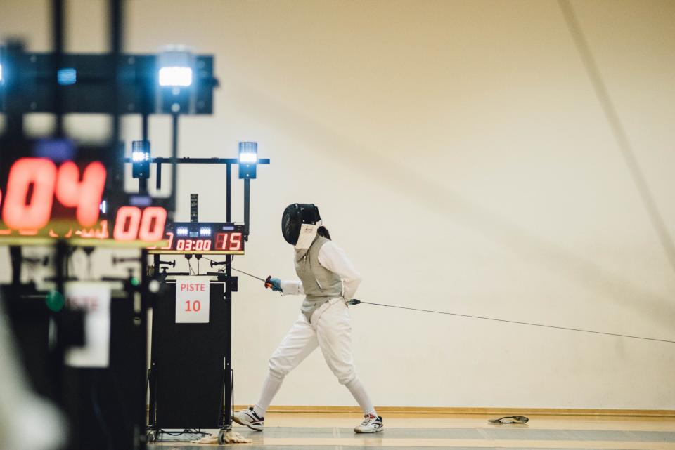 A fencer on a piste beside the electric scoring box, with its lamps and a red scoreboard.